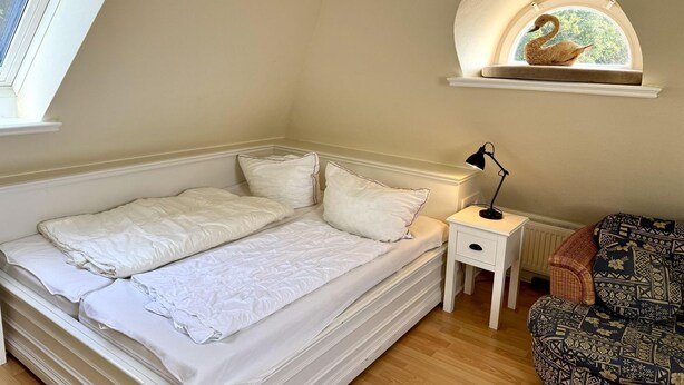 A bedroom with a bed, a nightstand, a lamp, a window, and a decorative swan on the wall. - Süderstraße 29a, Apt. 02 - 3-room upper-floor vacation apartment with a south-facing balcony for (Sylt-Ost)