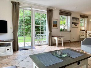 A room with a sliding glass door leading to a garden, a dining table with chairs, a television, and a small white cabinet. - Norderstraße, Apartment 1 - Cozy ground-floor vacation apartment with a fireplace and (Sylt)