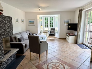 A living room with a sofa, armchair, TV, and a view of the outdoors through a large window. - Norderstraße, Apartment 1 - Cozy ground-floor vacation apartment with a fireplace and (Sylt)