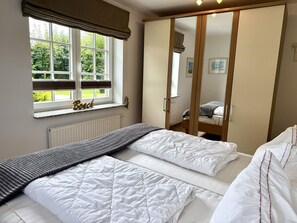 A bedroom with two beds, a window with a view of greenery, a radiator, and a wardrobe with a mirror. - Norderstraße, Apartment 1 - Cozy ground-floor vacation apartment with a fireplace and (Sylt)