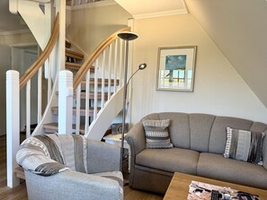 Ein Wohnzimmer mit einem grauen Sofa, gemusterten Kissen, einem Holzstuhl, einer Stehlampe und einer Treppe mit Holzbalken.