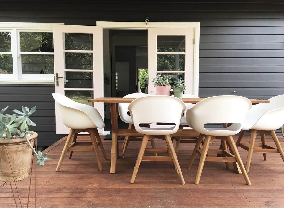 A patio with a wooden table and white chairs, a potted plant, and a door leading to a dark interior.