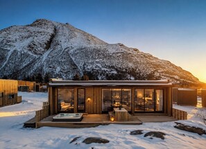 A modern cabin with a glass facade, wooden deck, and a hot tub.