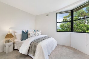 Ein Schlafzimmer mit einem Bett, einem Nachttisch, einer Lampe und einem Fenster mit Blick auf Bäume.