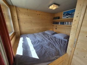 Ein kleines Zimmer aus Holz mit einem Bett, einem Fenster mit Vorhängen und einem Bücherregal.
