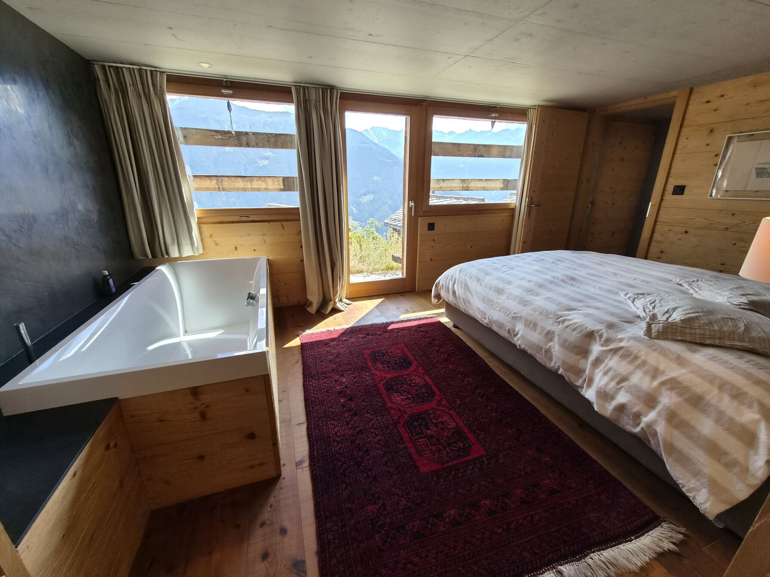 A bedroom with a wooden floor, a bed, a window with curtains, and a view of mountains.