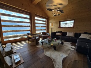 Ein gemütliches Blockhaus-Interieur mit einem Ledersofa, einem Glastisch und einem großen Fenster mit Blick auf die Berge.
