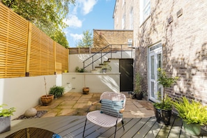 A courtyard with a wooden deck, a metal chair, potted plants, and a brick building with a staircase. - Beautiful 2BDR Home Right by Regents Park (Greater London)