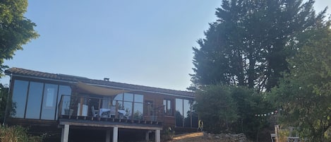 A modern house with a large deck, surrounded by trees and a clear sky.
