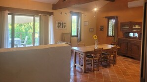 A dining area with a wooden table and chairs, a view of the outdoors, and a kitchen with wooden cabinets.