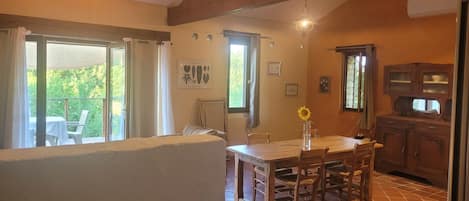 A dining area with a wooden table and chairs, a view of the outdoors, and a kitchen with wooden cabinets.