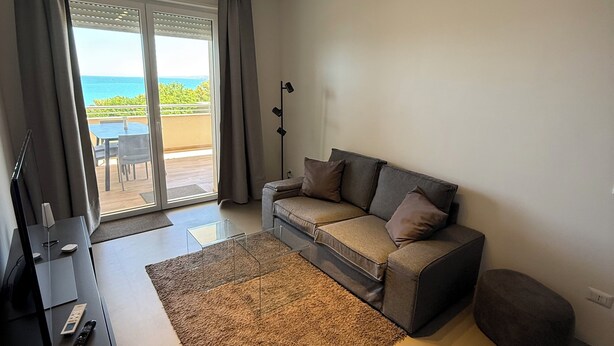 A modern living room with a sofa, a coffee table, and a TV stand. There is a large window with curtains, and a view of the outdoors. - Brand-New Sea View in Vasto Marina with Parking (Marina di Vasto)