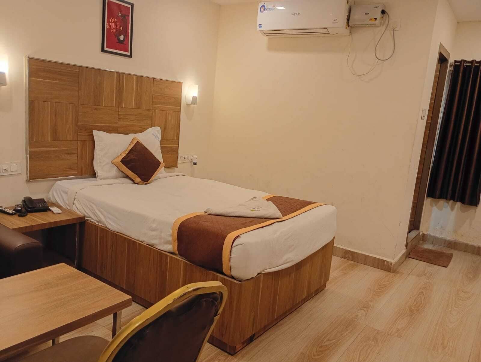 Een hotelkamer met een houten hoofdeinde, een bed met witte beddengoed en een bruin kussen, een nachtkastje met een telefoon, een stoel en een airconditioningunit aan de muur.