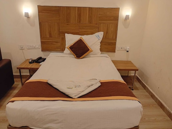 Quarto de hotel com cabeceira de madeira, cama com colcha marrom e branca, um travesseiro e mesa de cabeceira com telefone, além de mesa de apoio com luminária.