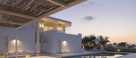 A modern outdoor pool area with a covered seating area and a stone wall in the background.