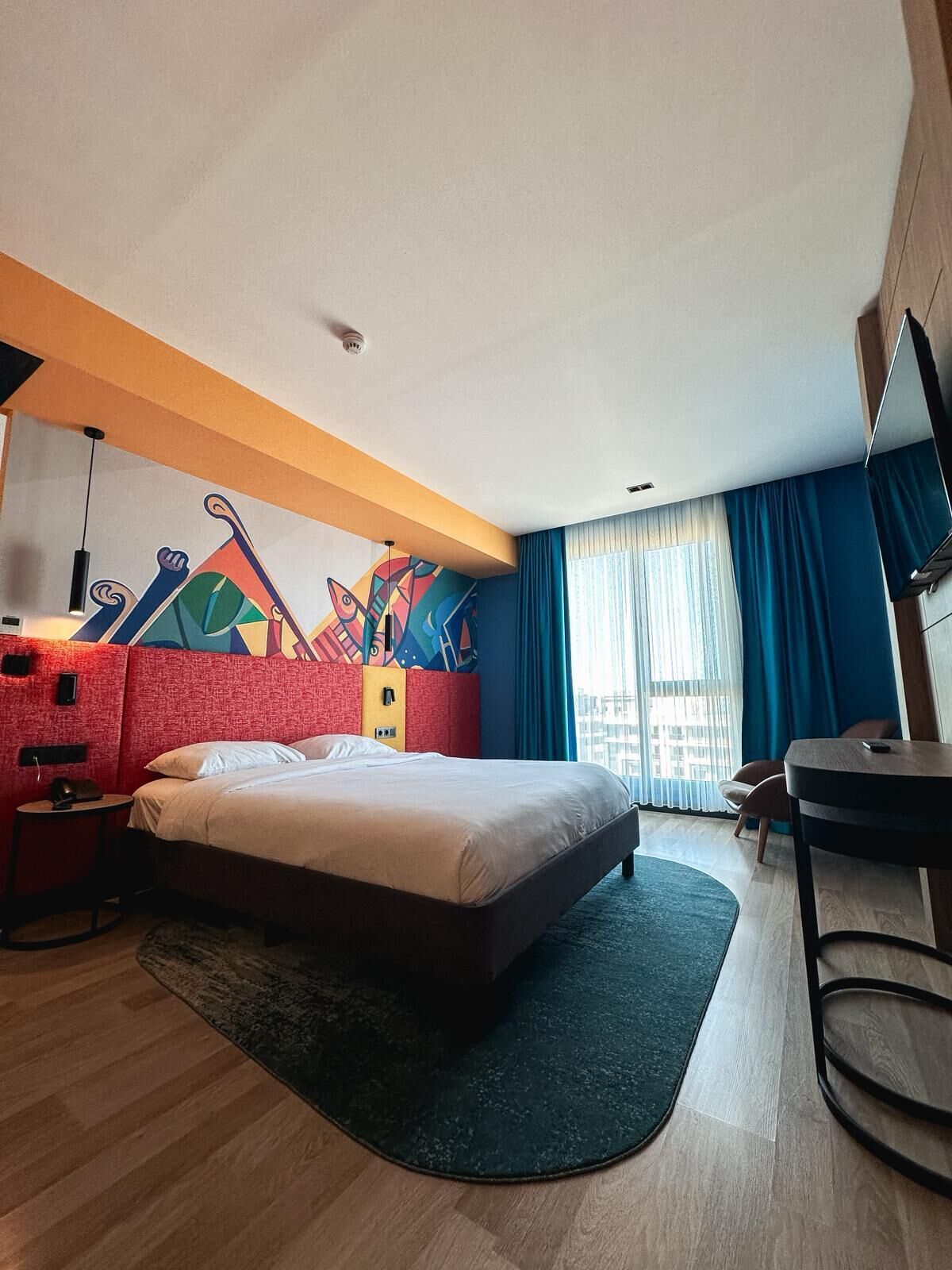 A modern hotel room with a large bed, a desk, a chair, a colorful abstract wall art, and a view of the city through the window.