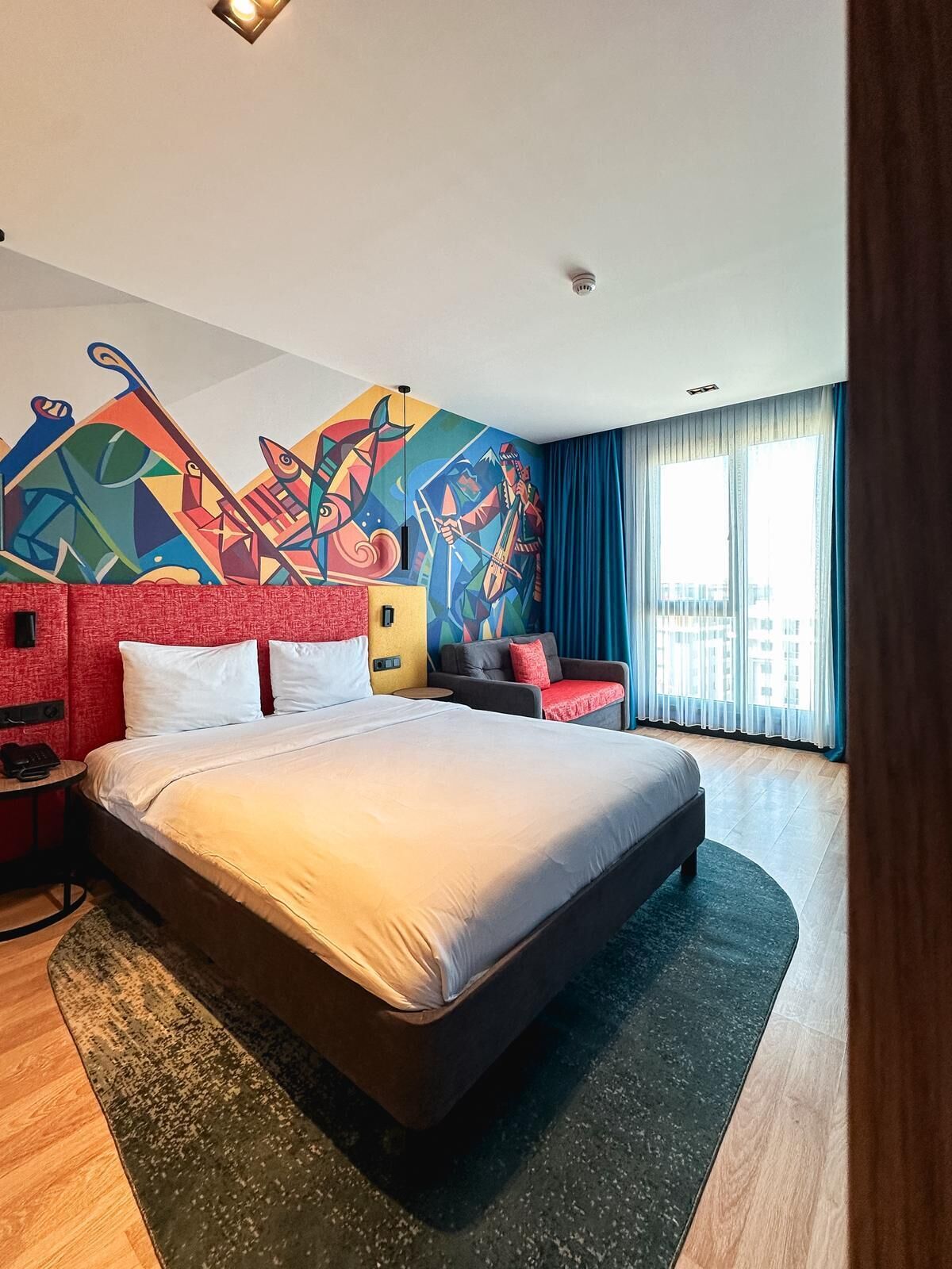 A hotel room with a large bed, a sofa, a colorful abstract mural, a window with curtains, and a small table with a phone.
