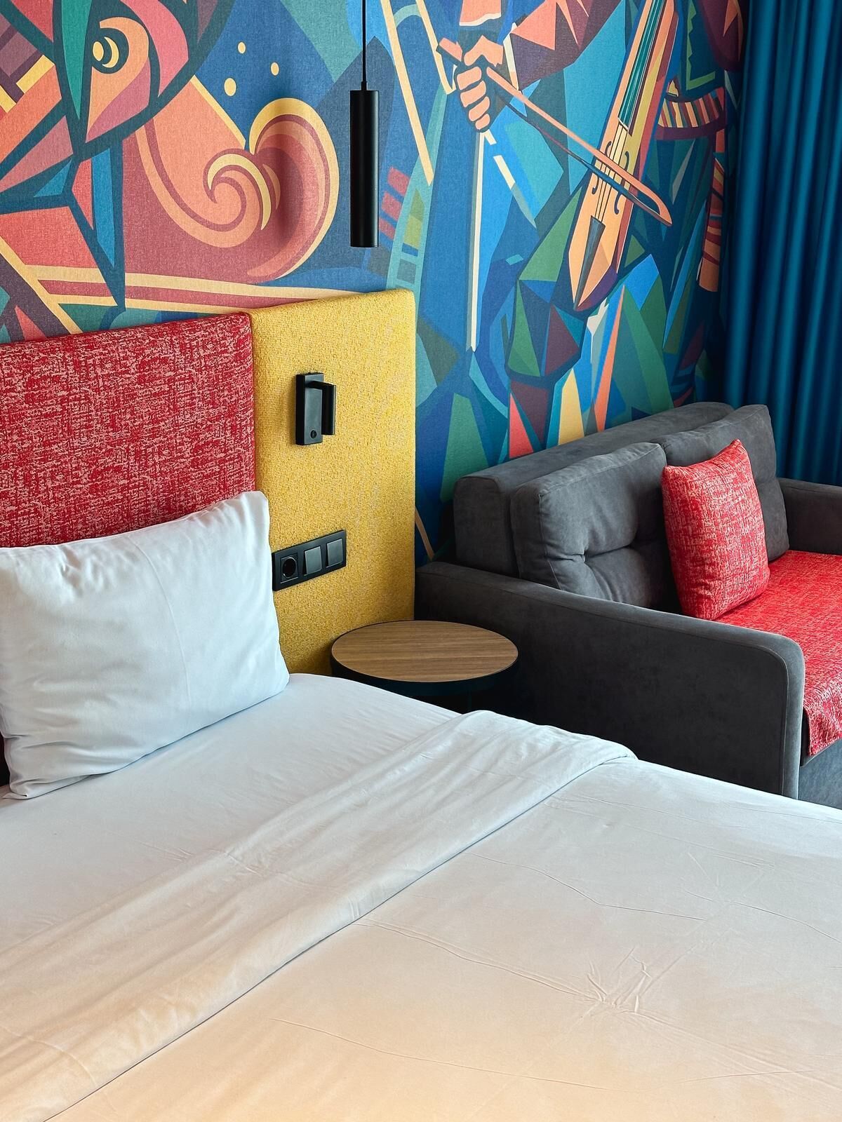 A hotel room with a bed, a sofa, a small round table, and a colorful abstract mural on the wall.