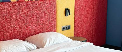 A hotel room with a bed, a red textured headboard, a colorful abstract mural on the wall, and a small table with a lamp.