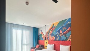 Una habitación de hotel con dos camas, un ventanal con cortinas y un mural abstracto colorido en la pared, además de una pequeña mesa con una silla.