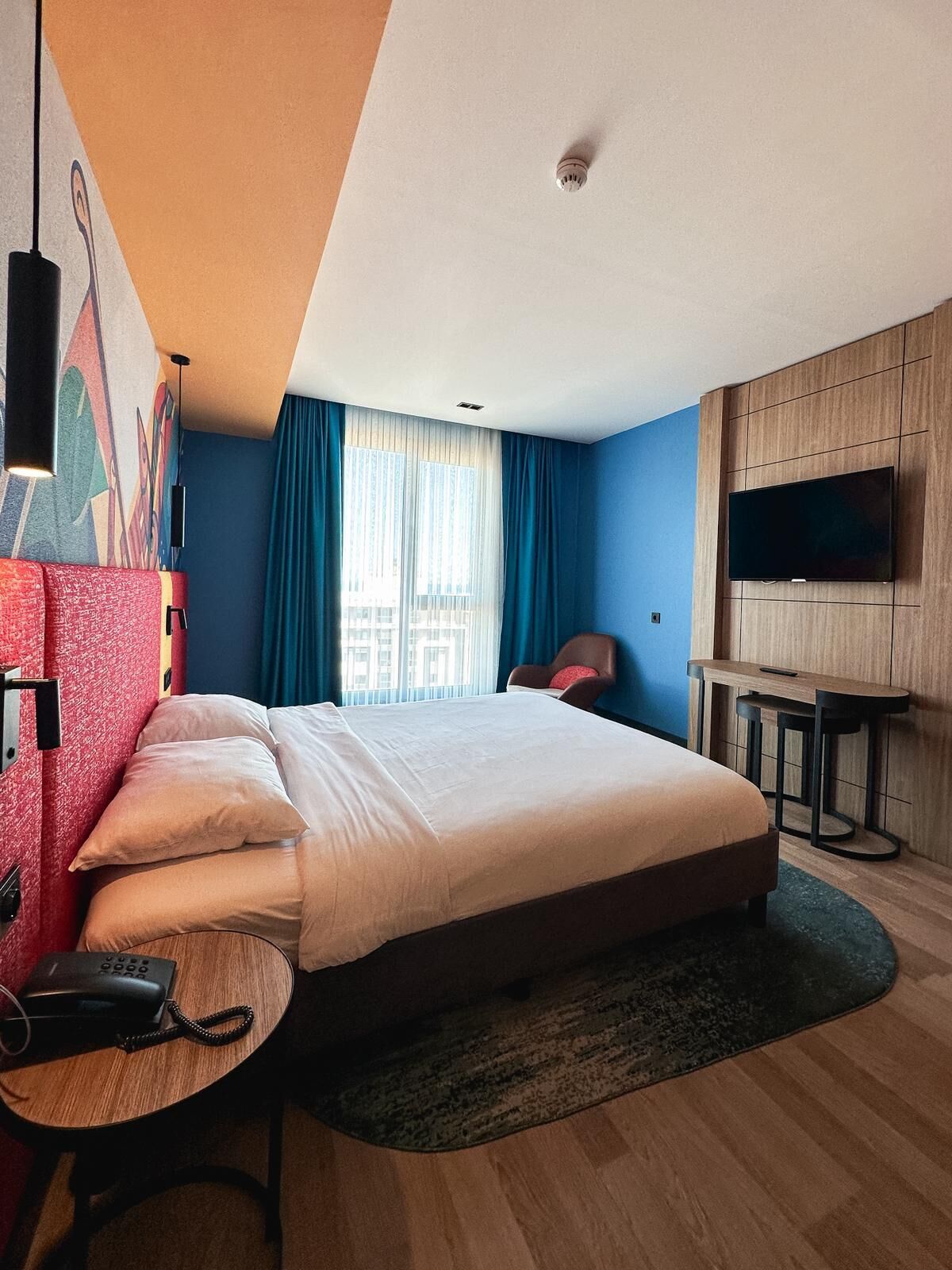 Habitación de hotel con una cama grande, un escritorio con teléfono, un televisor montado en la pared y una ventana con cortinas.