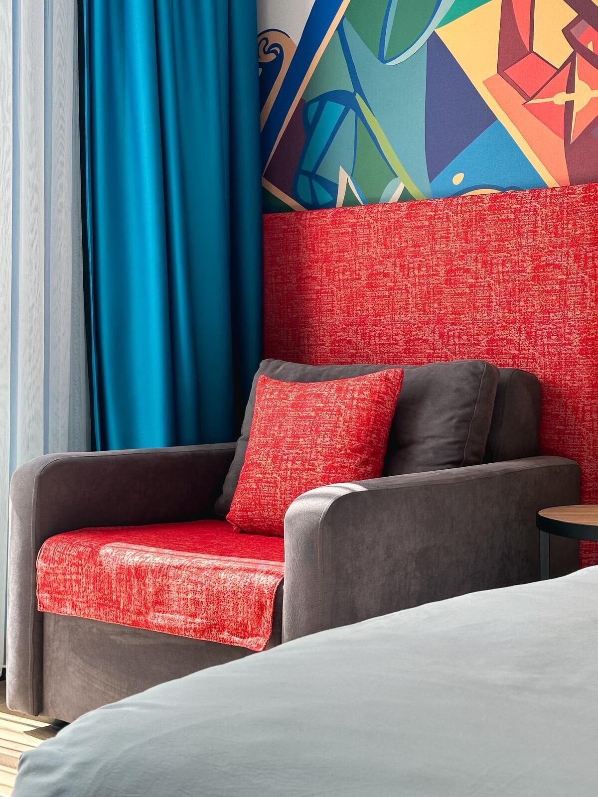 Quarto de hotel com um sofá cinzento, um cobertor vermelho estampado e uma peça de arte geométrica na parede.