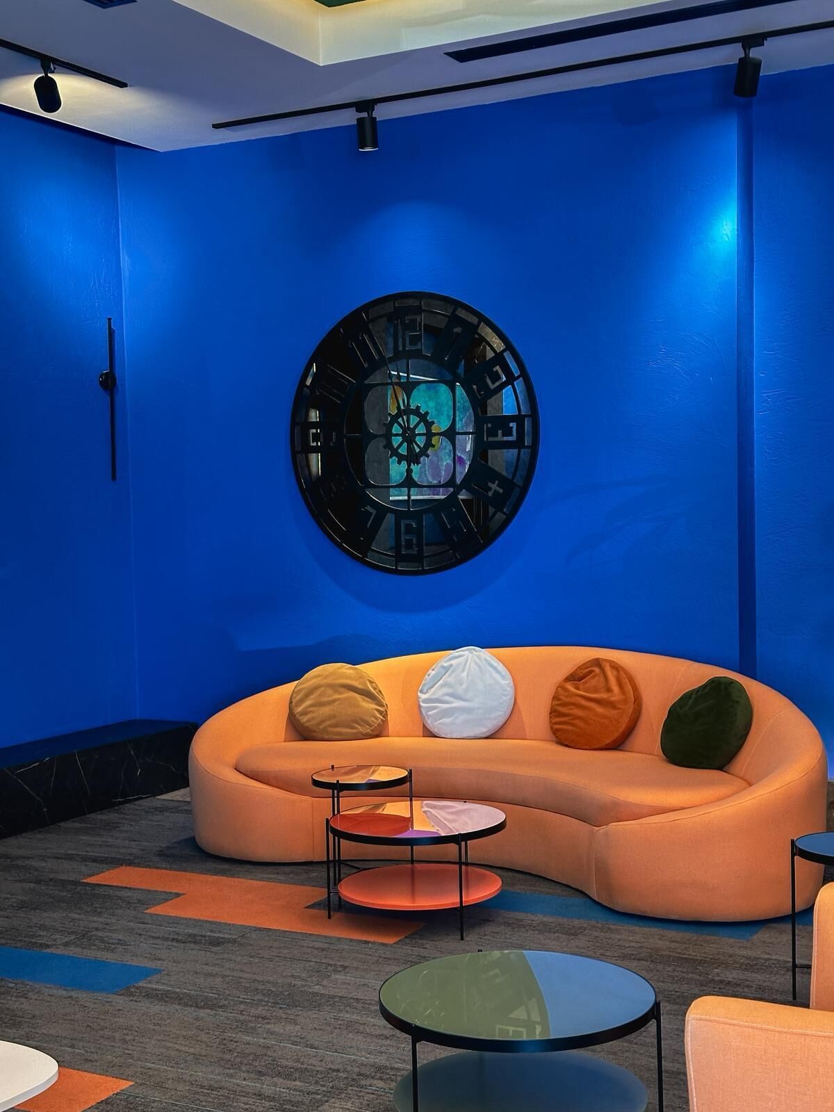 A modern interior with a blue wall, a round clock, a curved orange sofa, and small round tables.