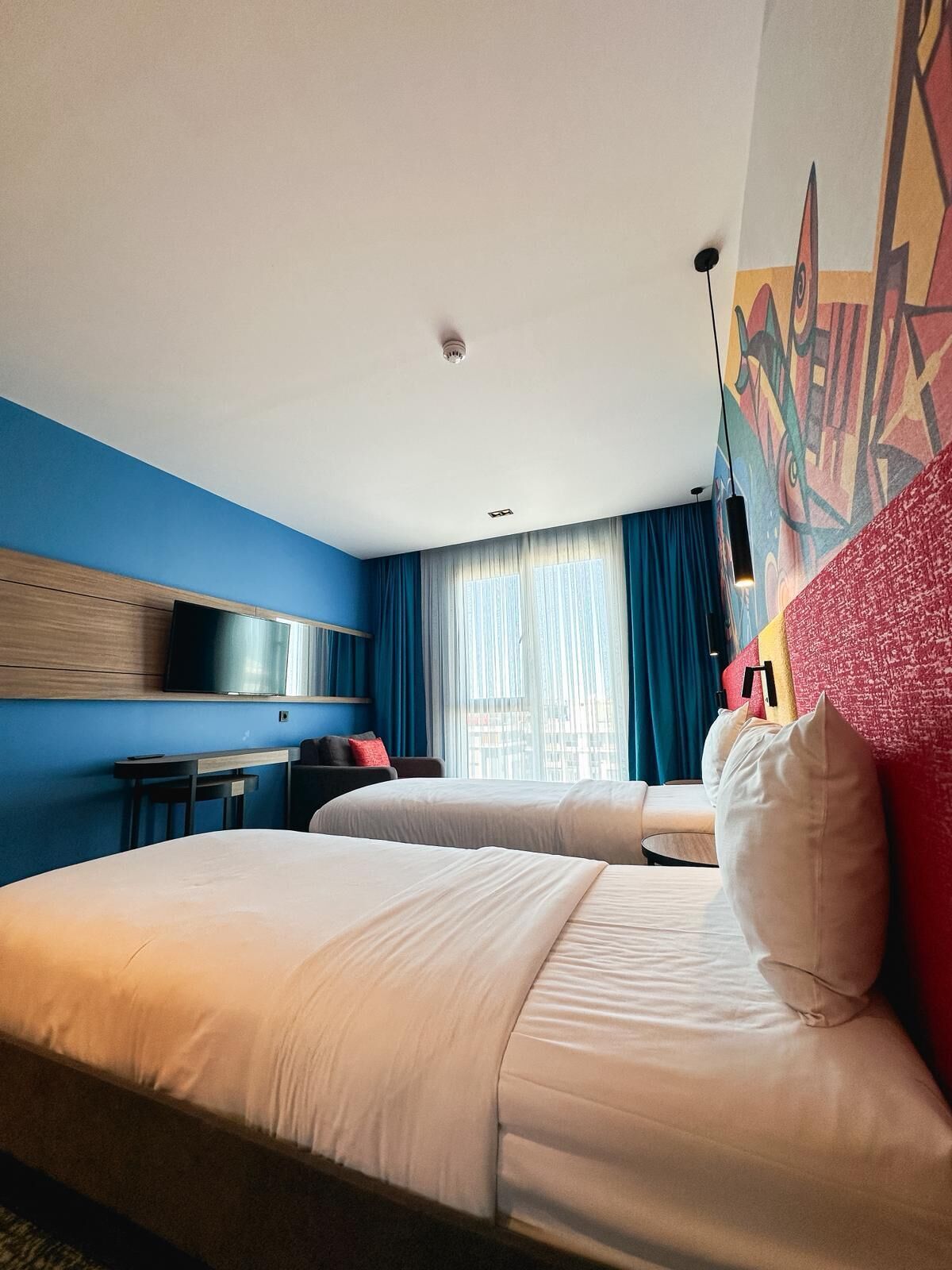 Une chambre d’hôtel avec un grand lit, un téléviseur fixé au mur, un bureau avec une chaise et une fenêtre avec des rideaux.