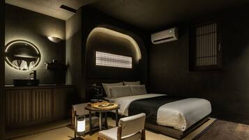 Een moderne hotelkamer met een bed, nachtkastje, stoel en een klein rond tafeltje met een lamp.