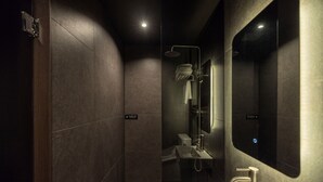 A modern bathroom with a dark tiled wall, a glass shower enclosure, a sink with a mirror, and a towel rack. - Cave Ryokan Hotel by Anook Seoul Jongno (Seoul)
