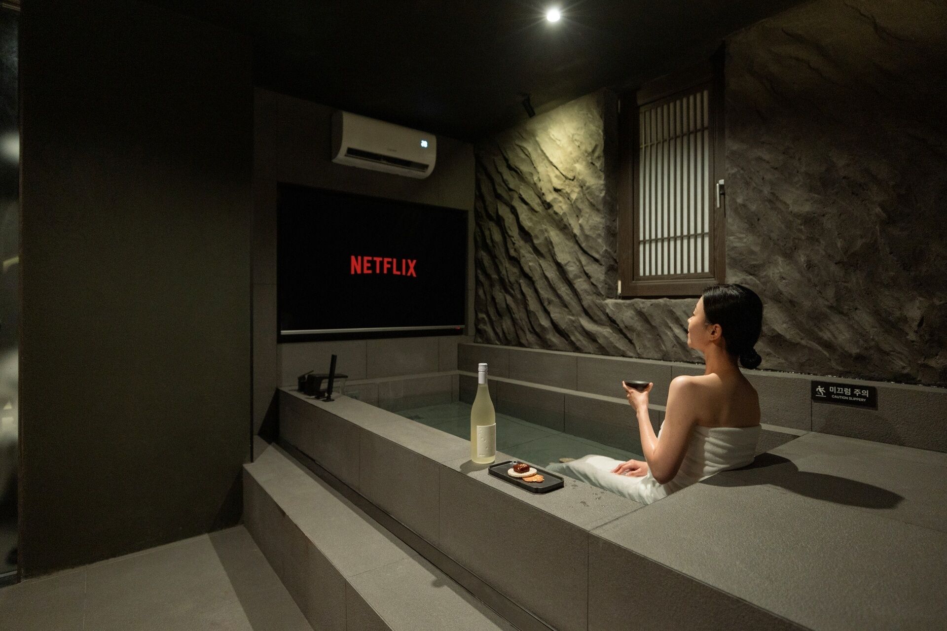 A person relaxing in a bathtub with a Netflix screen above, a bottle, and a tray with snacks.
