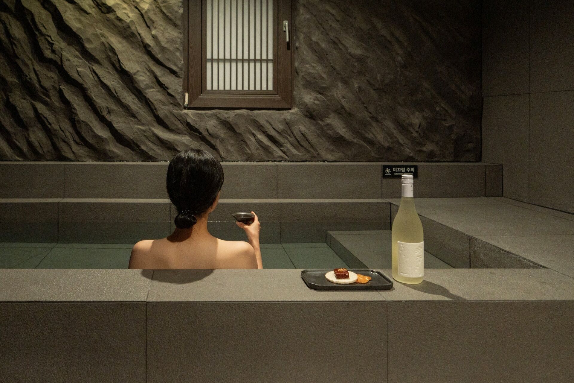 A person in a bathtub with a bottle of white wine and a tray with a dessert.