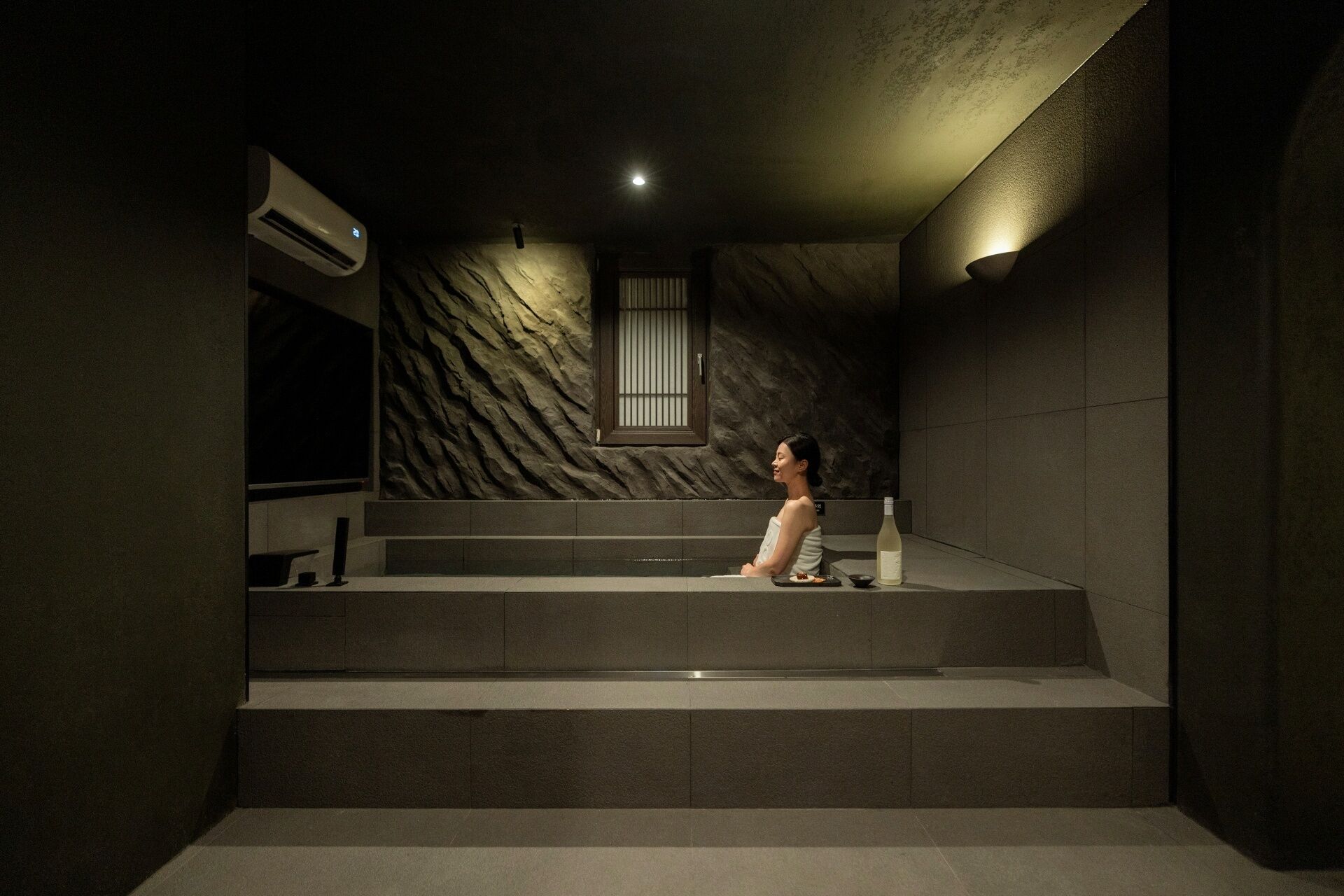 A person sitting on a staircase in a dimly lit room with a textured wall and a window.