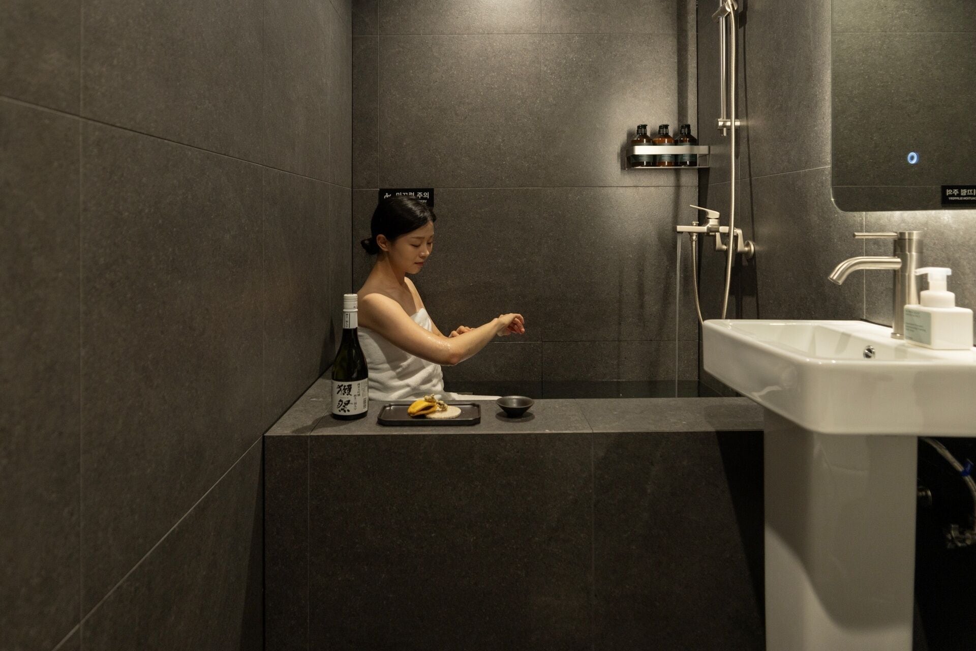 Une femme se trouve dans une salle de bain moderne, équipée d’un lavabo, d’un miroir et d’une douche.