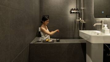 Une femme se trouve dans une salle de bain moderne, équipée d’un lavabo, d’un miroir et d’une douche.
