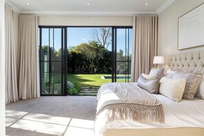 A bedroom with a large bed, a view of a garden through glass doors, and a ceiling with recessed lighting. - Belle Grove (Cape Town)