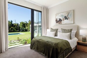 A bedroom with a large bed, a painting, a nightstand, and a view of a pool and greenery. - Belle Grove (Cape Town)