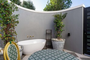 Een moderne badkamer in de open lucht met een vrijstaand bad, een klein plankje en twee potten met planten.