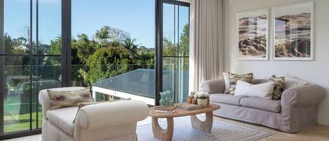 A spacious living room with a large glass balcony, a beige sofa set, a wooden coffee table, and two framed landscape paintings.