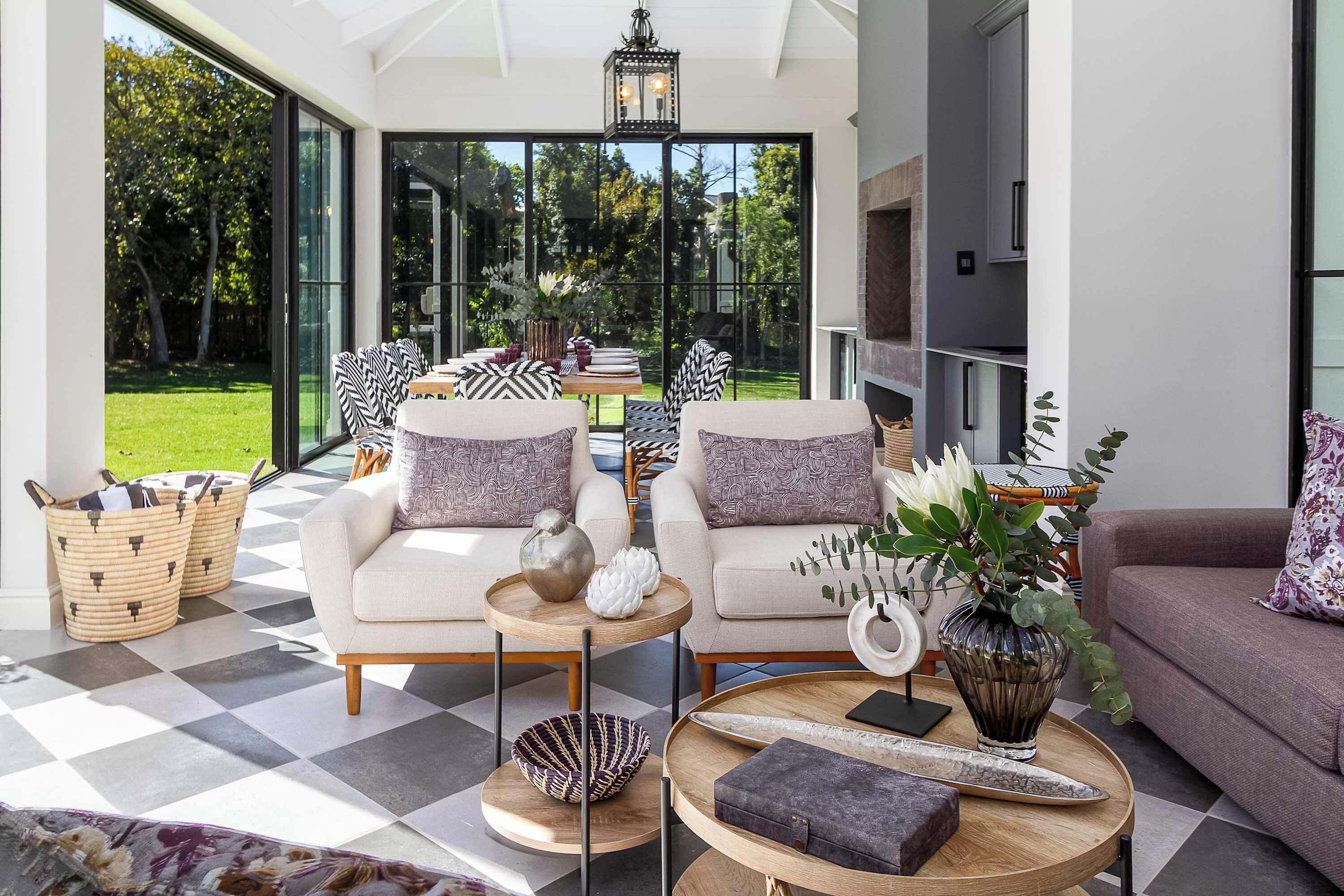 Una sala de estar moderna con piso a cuadros, dos sofás con cojines estampados, una mesa de centro con objetos decorativos y vistas a un jardín a través de amplias ventanas.