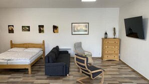 A bedroom with a wooden bed, a dark sofa, a chair, a dresser, and wall-mounted artwork. - Apartmány Statek Bezdědice (Hostomice / Středočeský kraj)