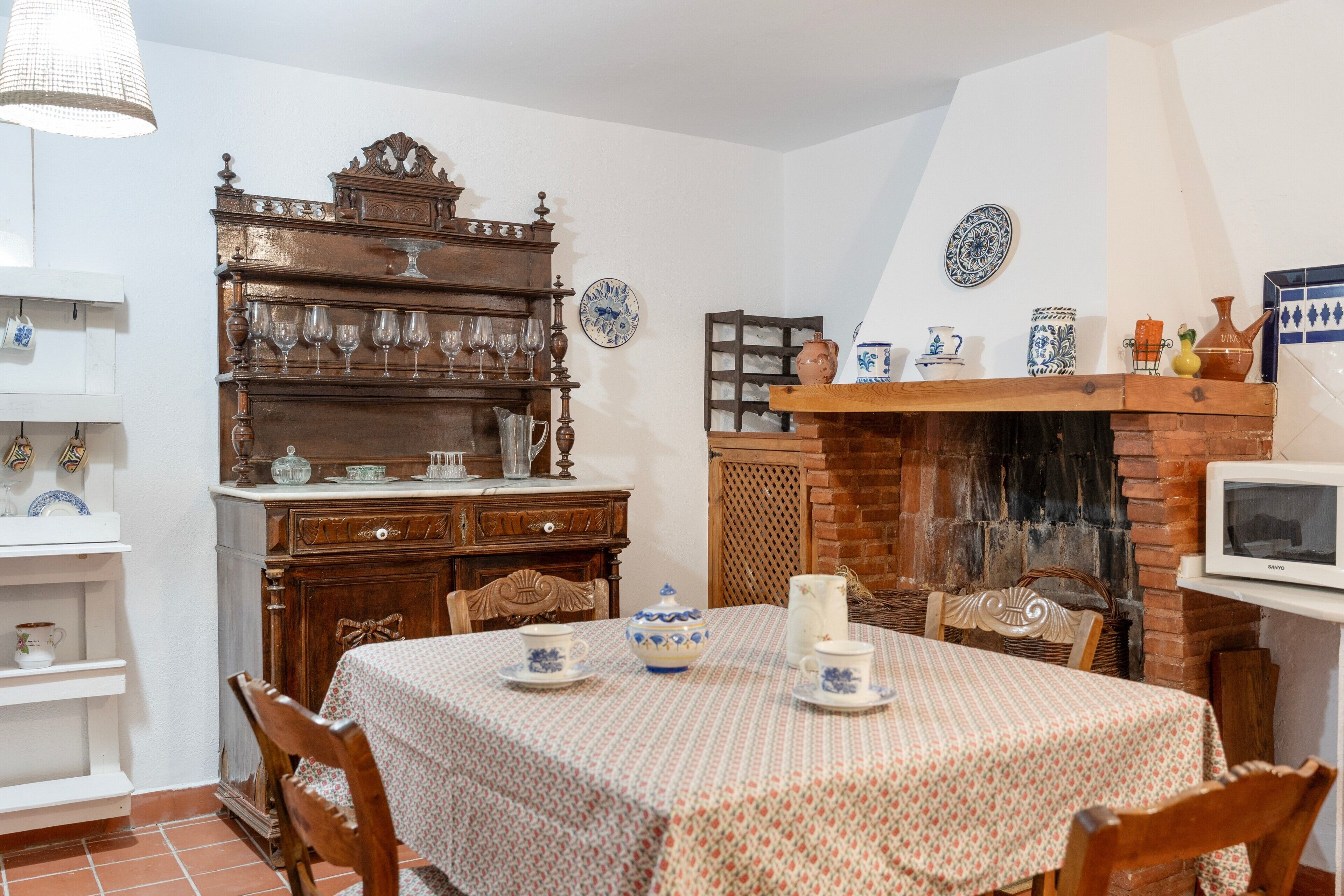 Un comedor con una mesa dispuesta para cuatro personas, rodeada de sillas. Un armario de madera con cristalería se encuentra a la izquierda, y una chimenea de ladrillo con un televisor a la derecha.