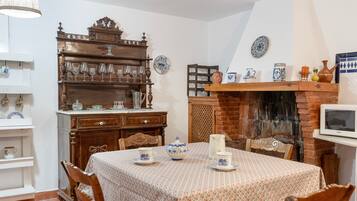 Un comedor con una mesa dispuesta para cuatro personas, rodeada de sillas. Un armario de madera con cristalería se encuentra a la izquierda, y una chimenea de ladrillo con un televisor a la derecha.