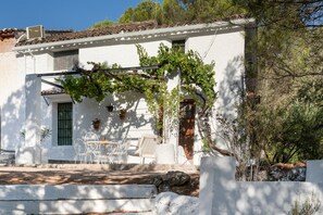 A white building with a tiled roof, a pergola with vines, outdoor seating, and a stone wall.