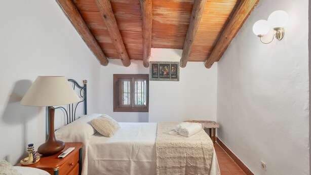 A bedroom with a wooden ceiling, a bed with a white bedspread, a bedside table with a lamp, and a small window.