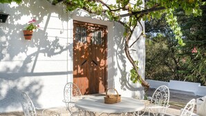 A white-walled building with a wooden door, white metal chairs, and a round table with a basket.