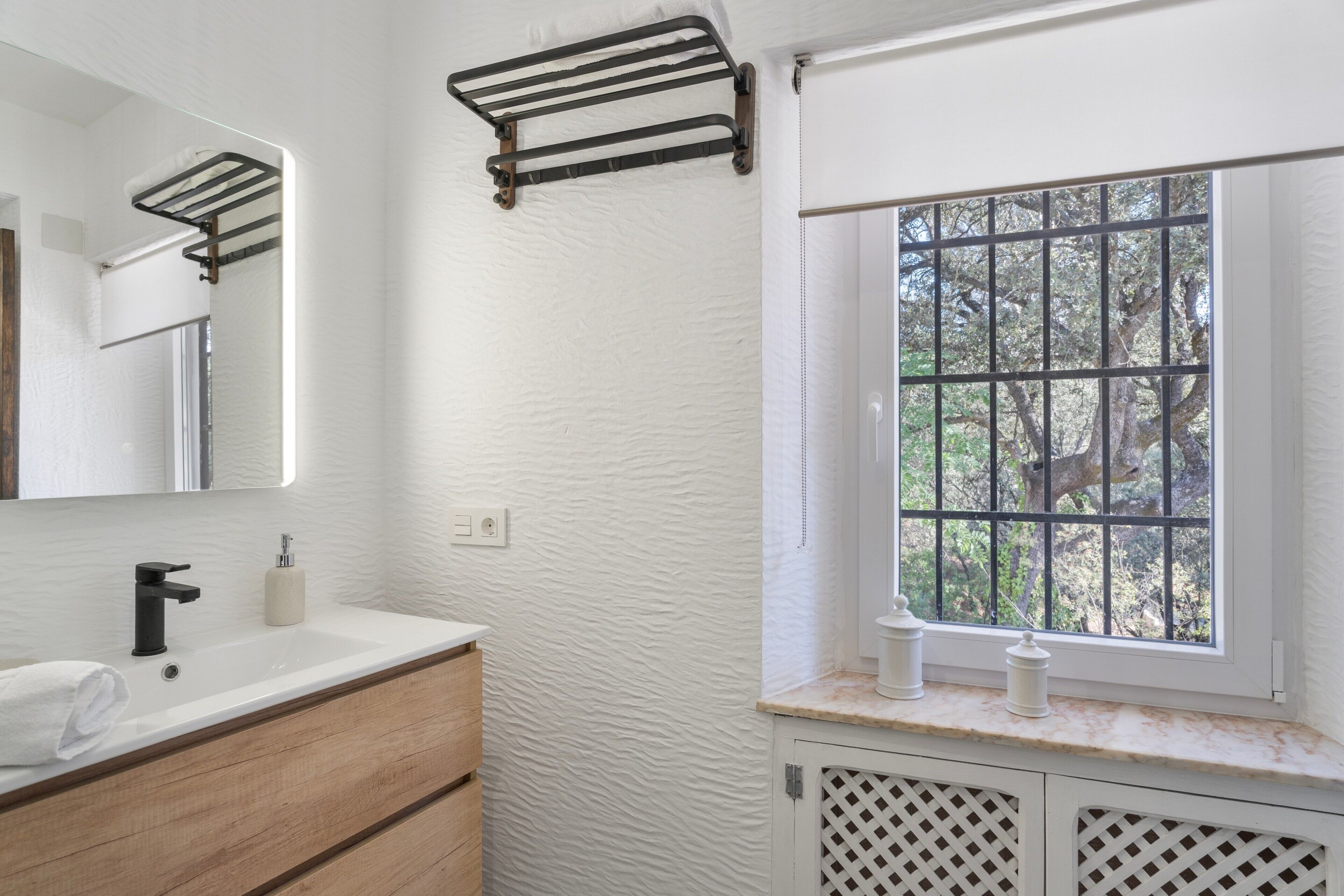 Un baño con pared blanca texturizada, tocador de madera, grifo negro, toallero, ventana con barrotes y espejo.