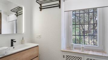 Un baño con pared blanca texturizada, tocador de madera, grifo negro, toallero, ventana con barrotes y espejo.