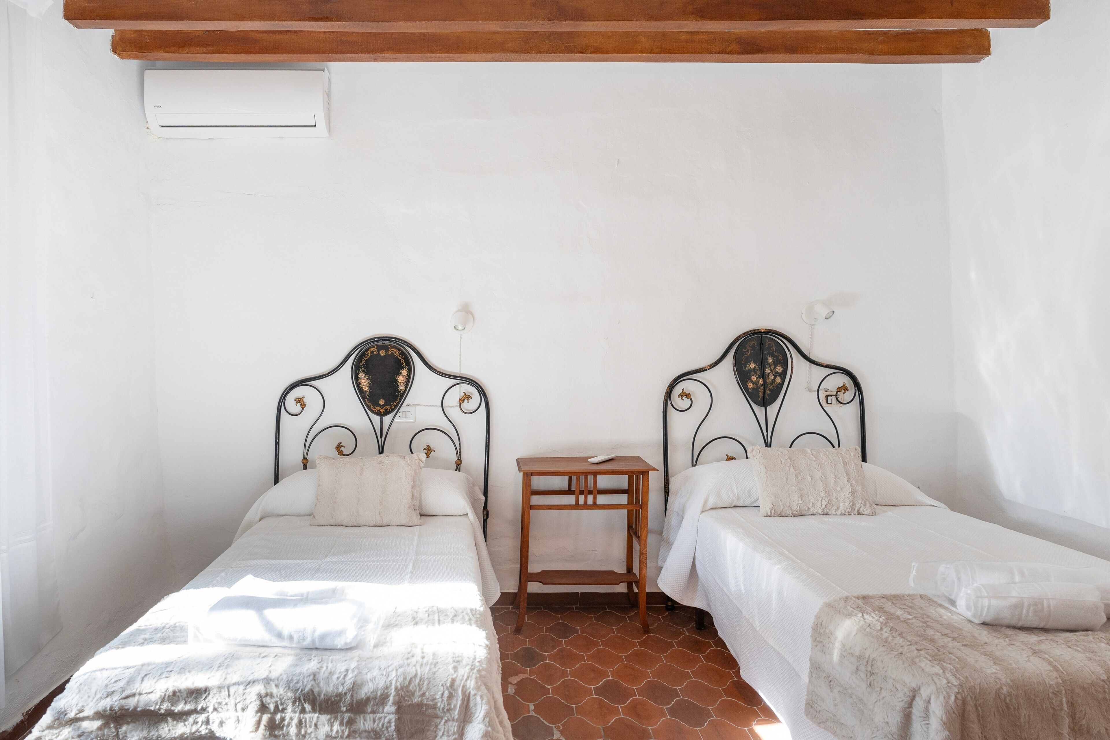 A room with two beds, a small wooden table, and a white ceiling with a vent.