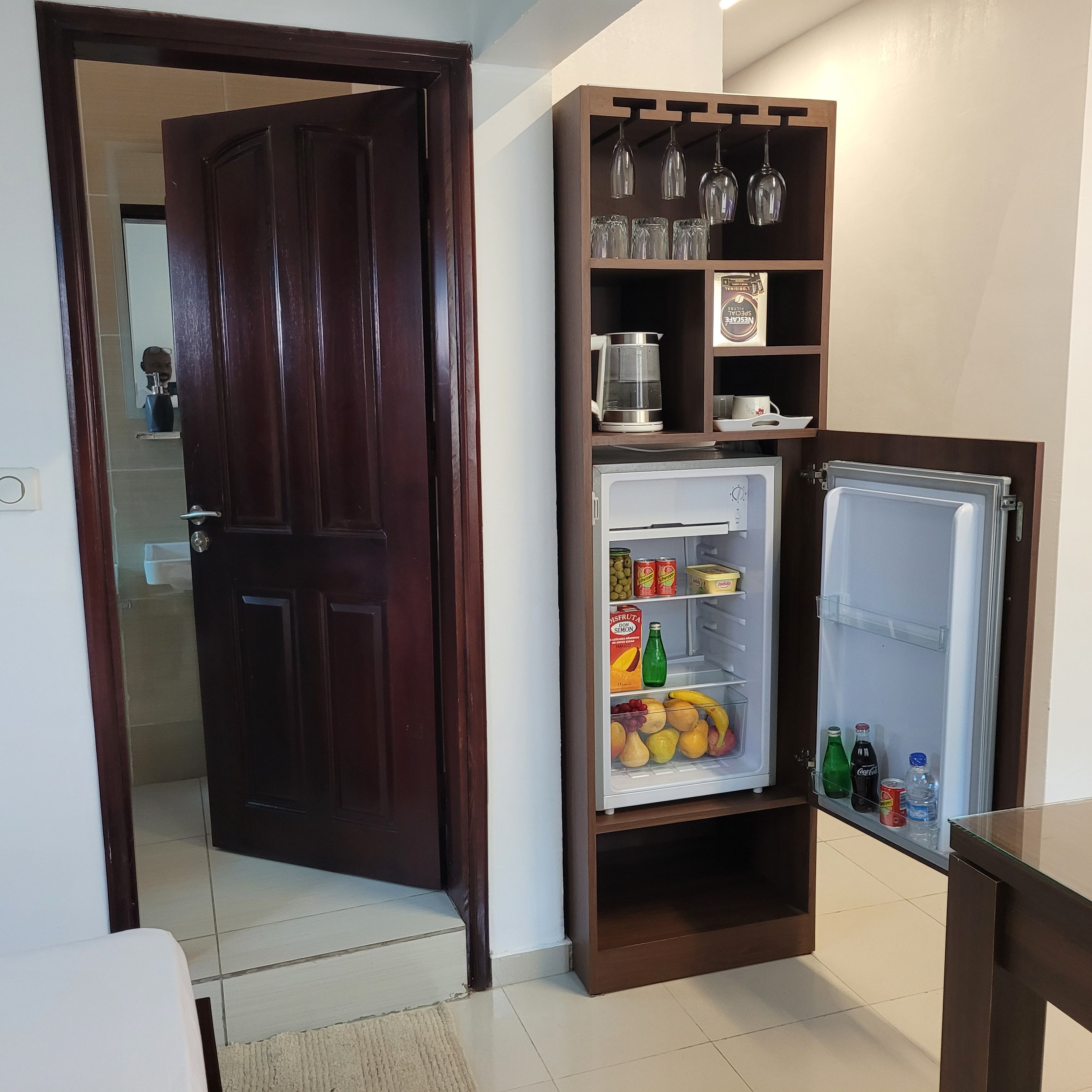 Una cocina compacta con refrigerador, cafetera y un aparador de vidrio con copas de vino.